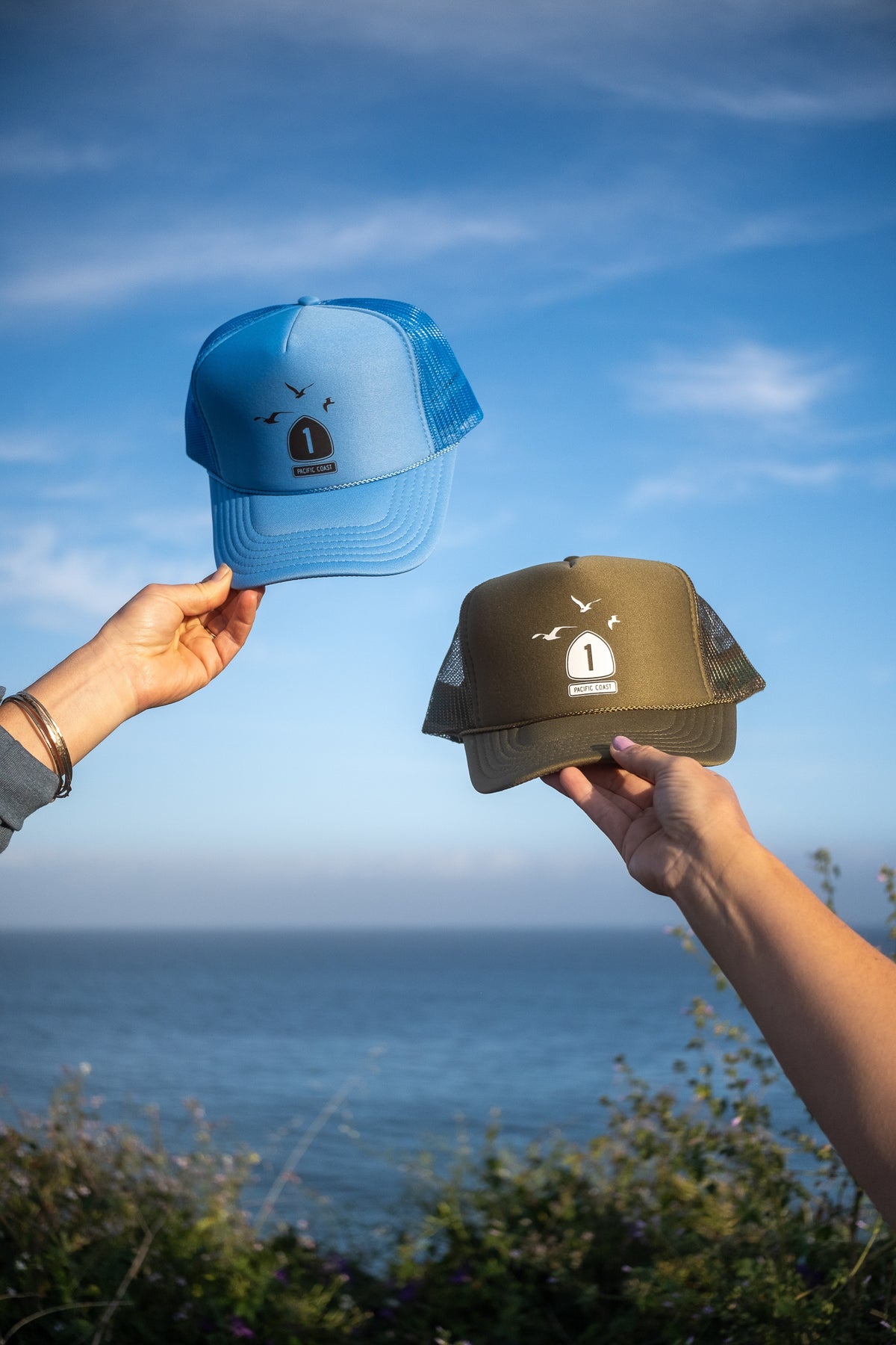 Two trucker hats are held up by hands against an outdoor coastal background. One hat is light blue and the other is olive green, both featuring a front logo patch with a bird and the number 1.
