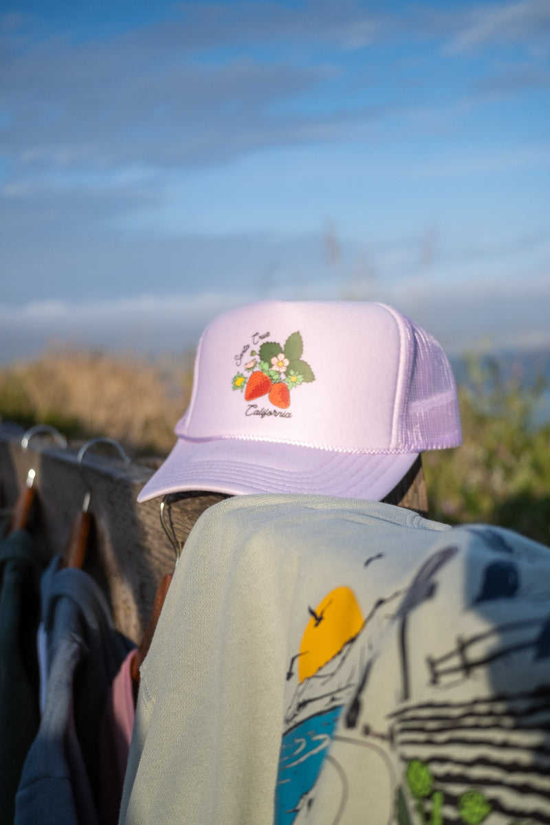A light purple/lavender trucker hat with a strawberry graphic and the text 'Santa Cruz California.'