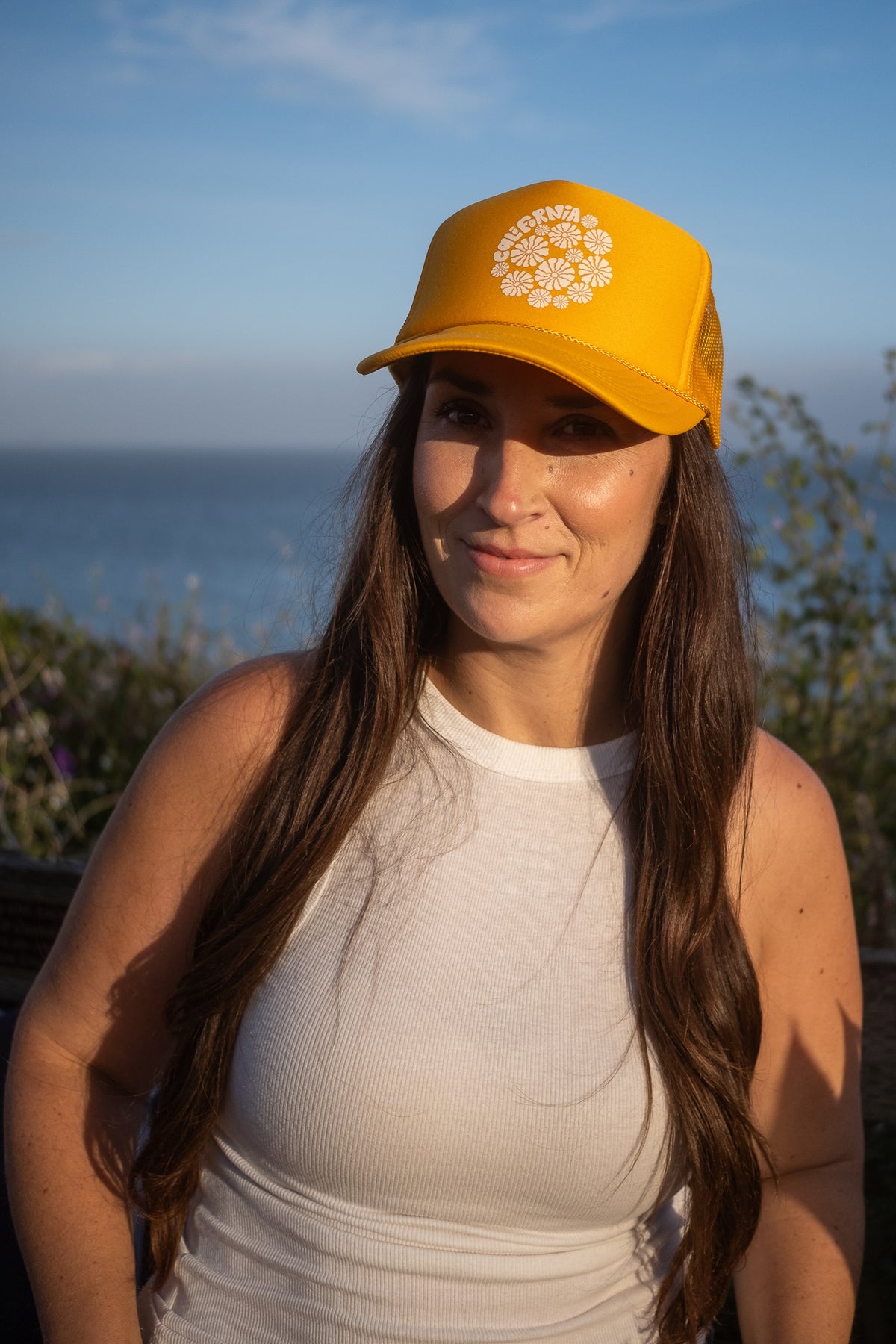 A woman with long brown hair wearing a yellow baseball cap with a white floral design and the text 'SUNRISE', paired with a white ribbed tank top, photographed outdoors with a scenic ocean background.