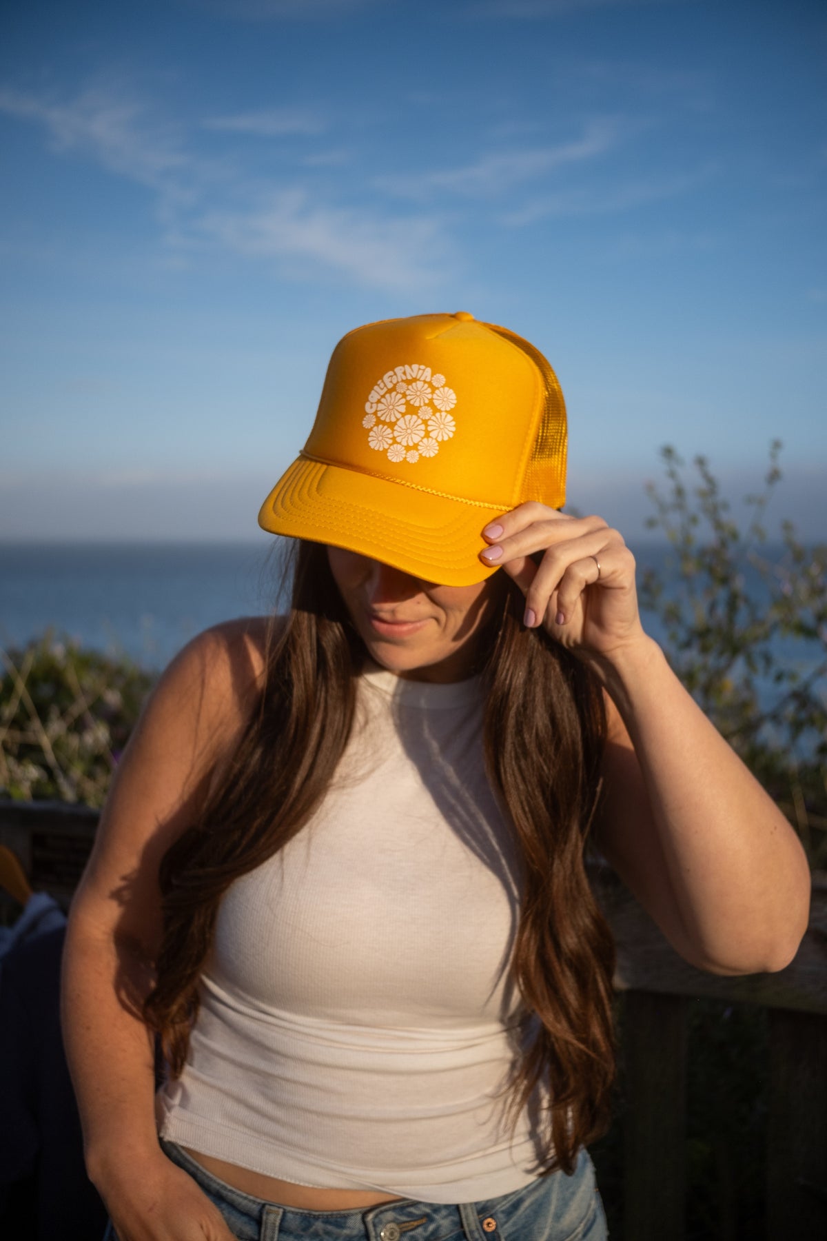 A woman outdoors wearing a bright yellow trucker hat with a white circular floral emblem on the front, holding the brim over her eyes. The background shows a blurred blue sky and ocean.