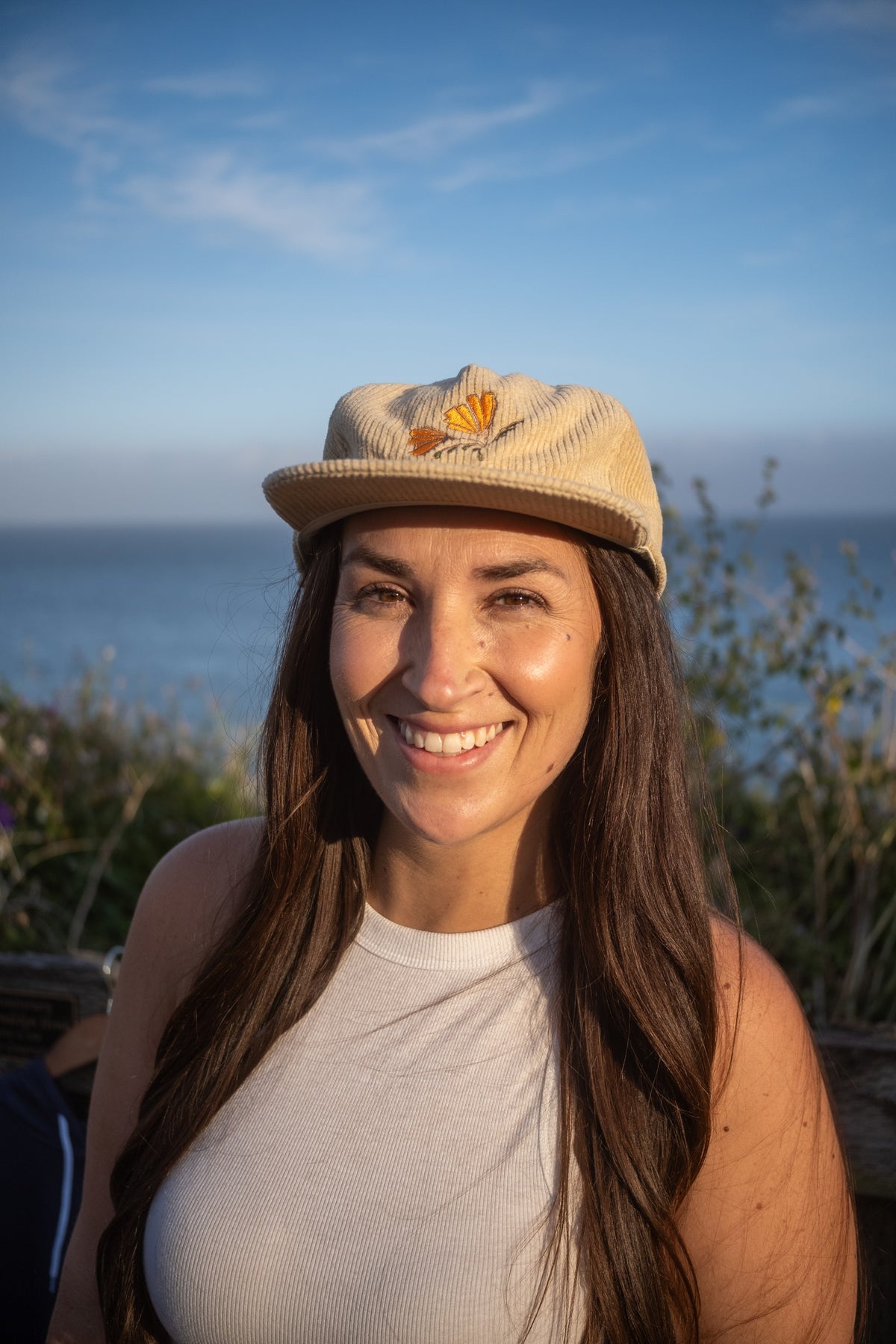A smiling woman outdoors wearing a beige corduroy cap with a small orange flower detail and a white sleeveless top, with ocean and sky in the background.