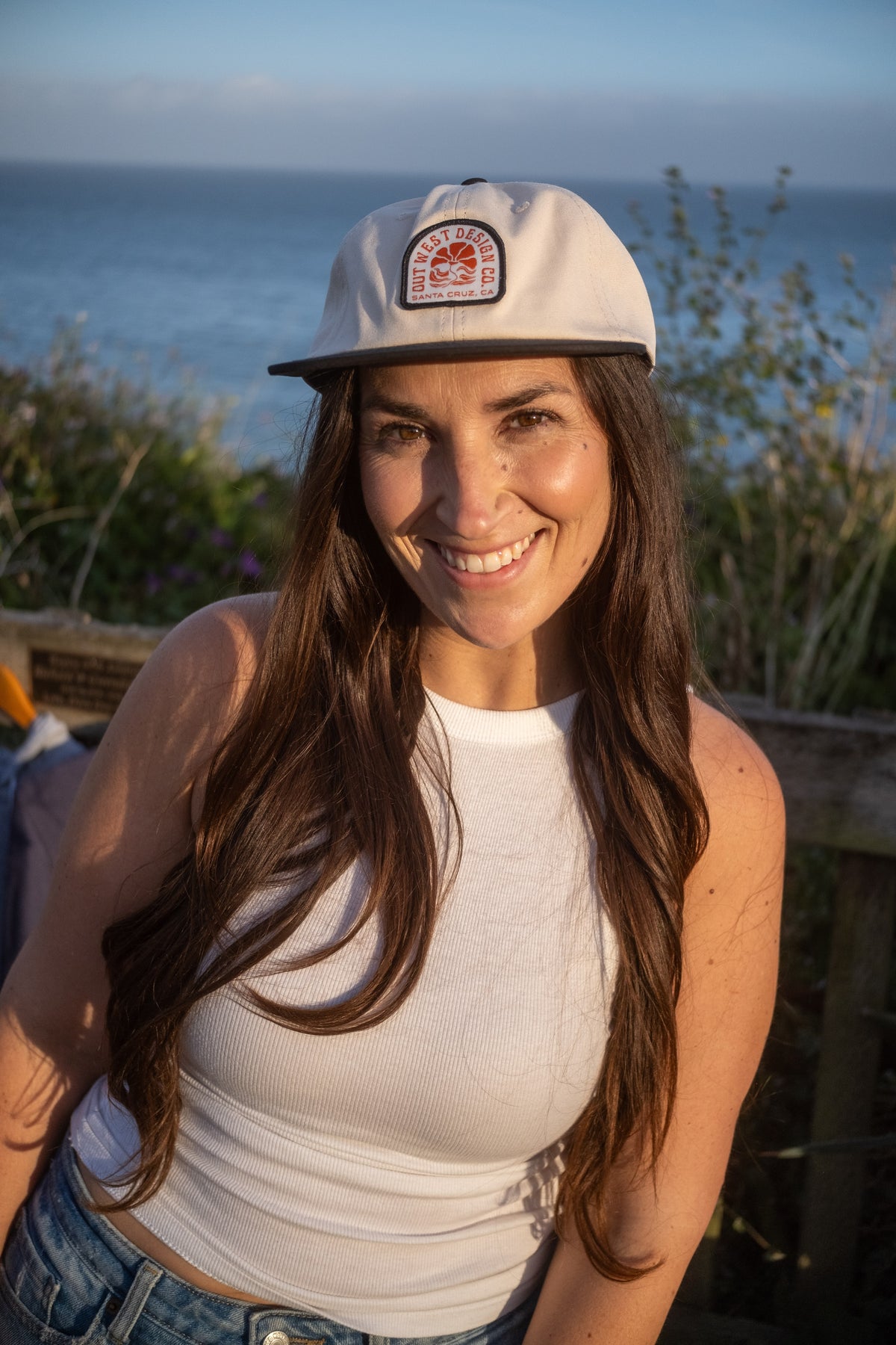 A smiling woman outdoors by the ocean wearing a light beige baseball cap with a circular orange and white logo patch on the front, a white sleeveless top, and blue jeans.