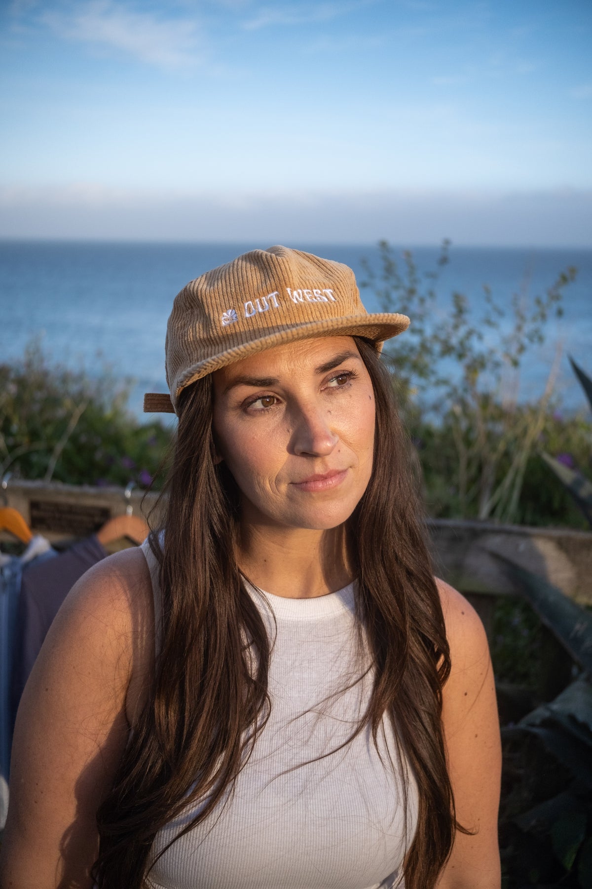 A woman with long brown hair wearing a beige corduroy cap with 'OUT WEST' embroidered on the front, set against a coastal background with the ocean and sky.