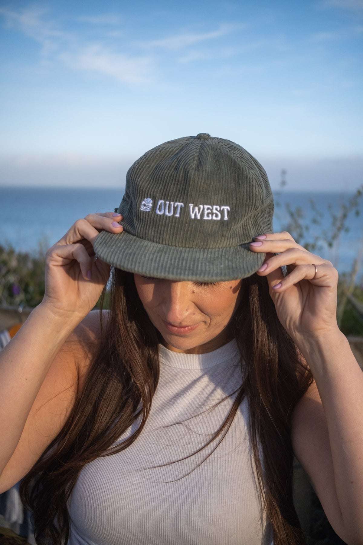 A woman wearing an olive green corduroy baseball cap with 'OUT WEST' embroidered in white on the front, holding the brim in front of an outdoor coastal background.