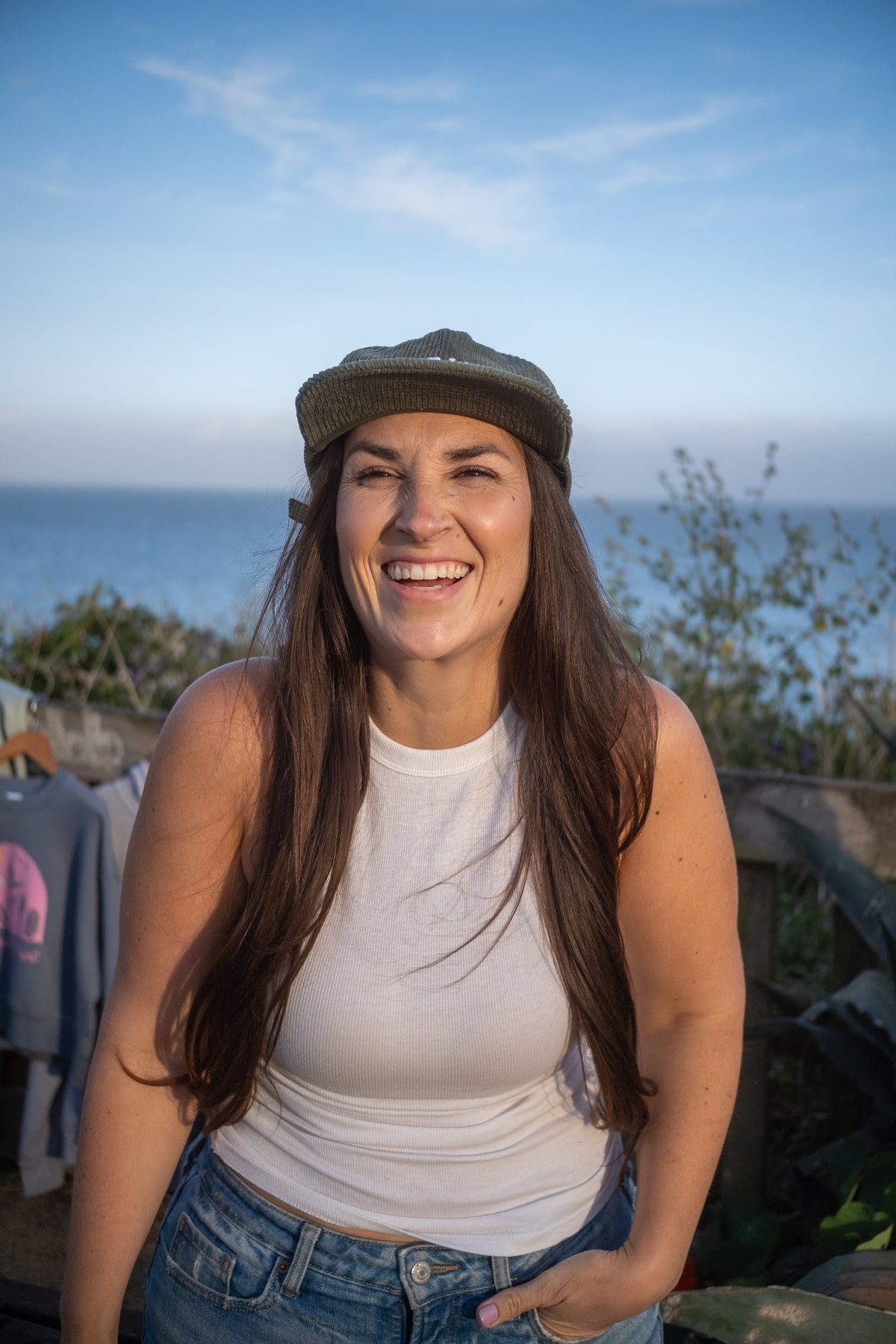 A smiling woman with long brown hair wearing a white tank top, blue jeans, and an olive green cap, standing outdoors with the ocean and sky in the background.