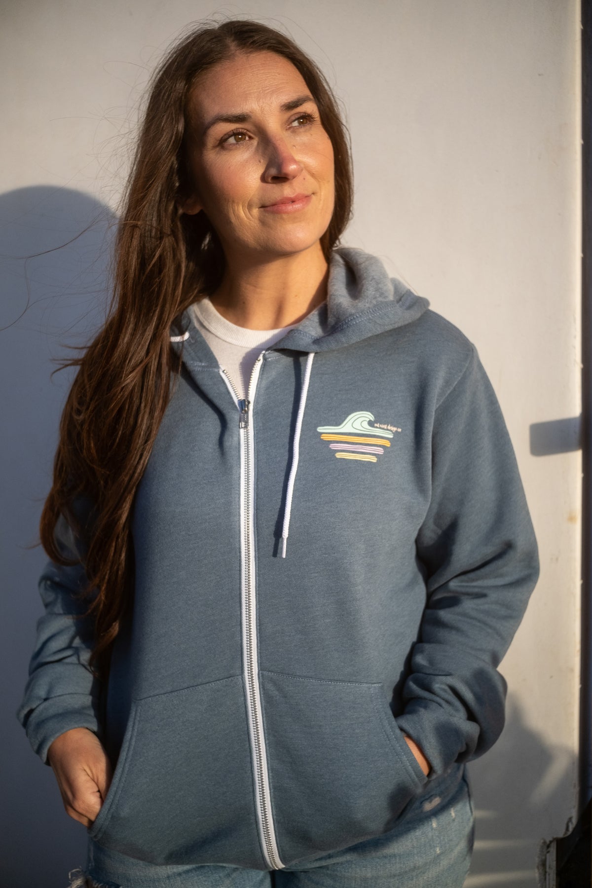 A woman with long brown hair wearing a slate blue zip-up hoodie with a white zipper and a small multicolor wave graphic on the left chest, standing against a white wall.