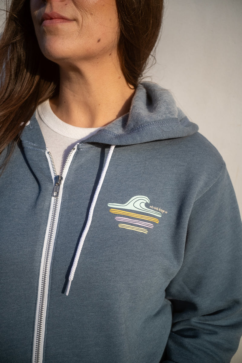 A close-up of a person wearing a heathered slate blue zip-up hoodie with a white drawstring and a small multicolor wave graphic on the left chest, over a white t-shirt.