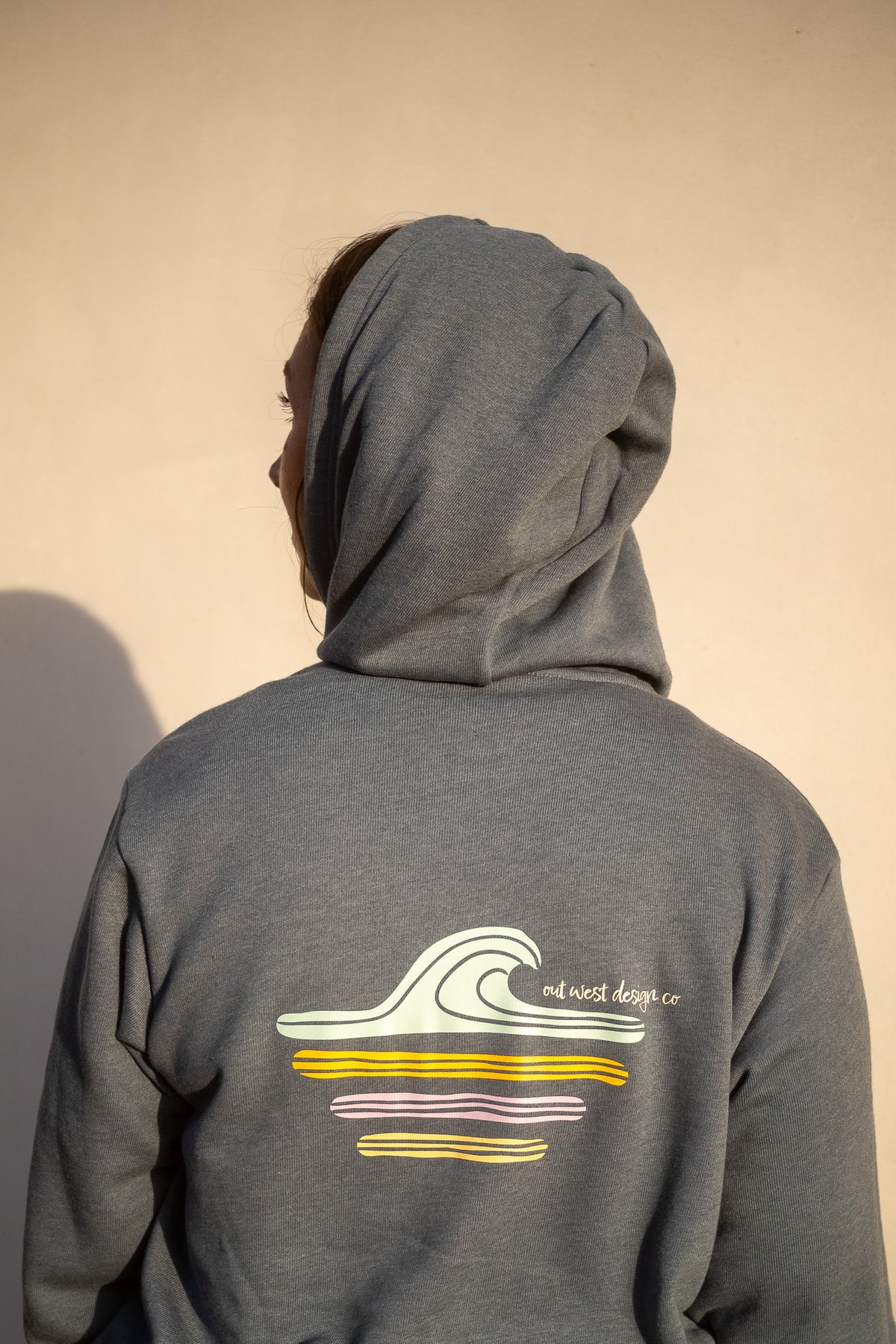 A person shown from behind wearing a heather gray hoodie with a graphic of a white wave and yellow and purple stripes on the back, set against a plain beige wall.