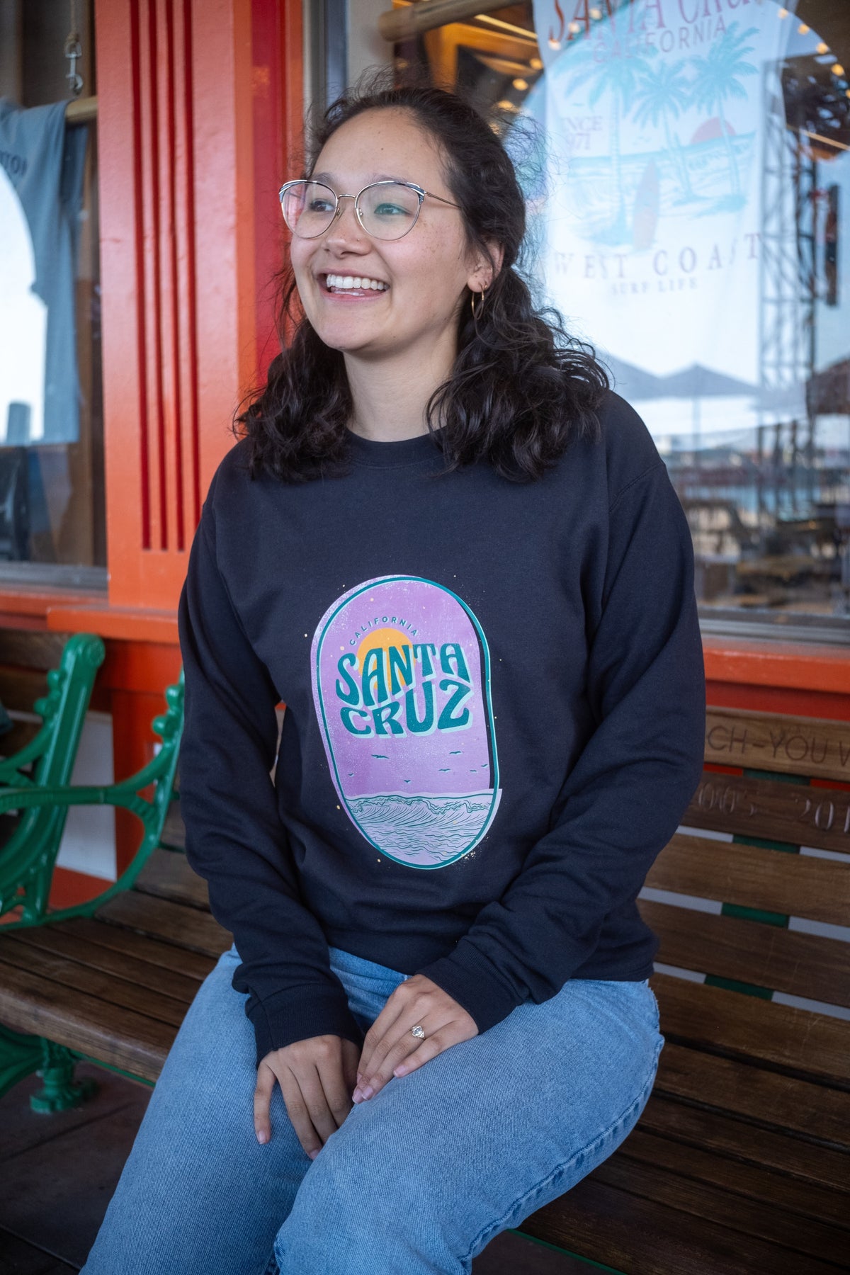 A woman with glasses and dark hair sits smiling on a wooden bench, wearing a black crewneck sweatshirt with a colorful 'Santa Cruz' graphic on the front and blue jeans.