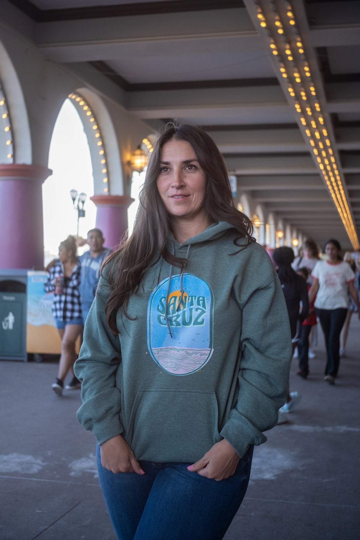 A woman wearing a sage green hoodie with a 'Santa Cruz' graphic on the front, standing in a covered outdoor walkway with people and architectural columns in the background.