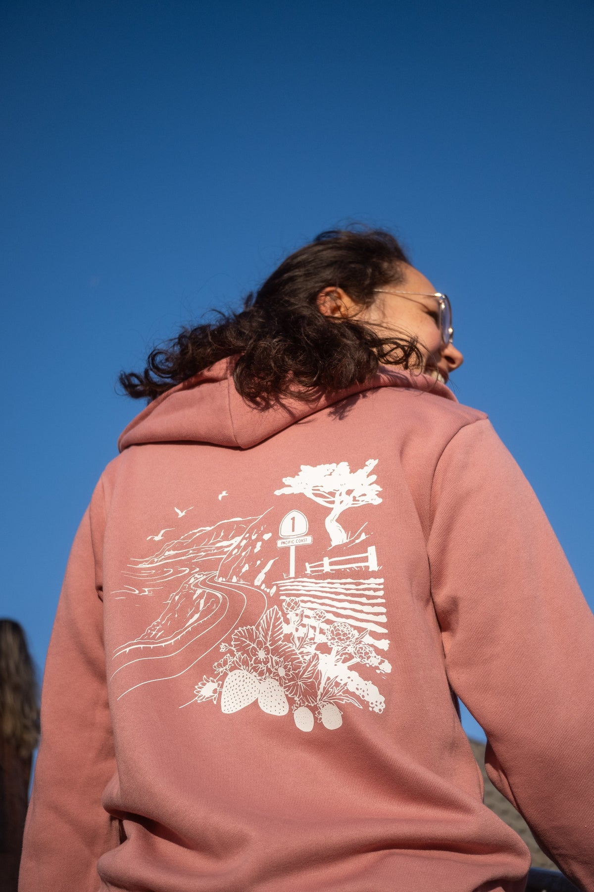 A person outdoors wearing a dusty rose pink hoodie with a large white graphic print on the back, photographed from behind against a clear blue sky.