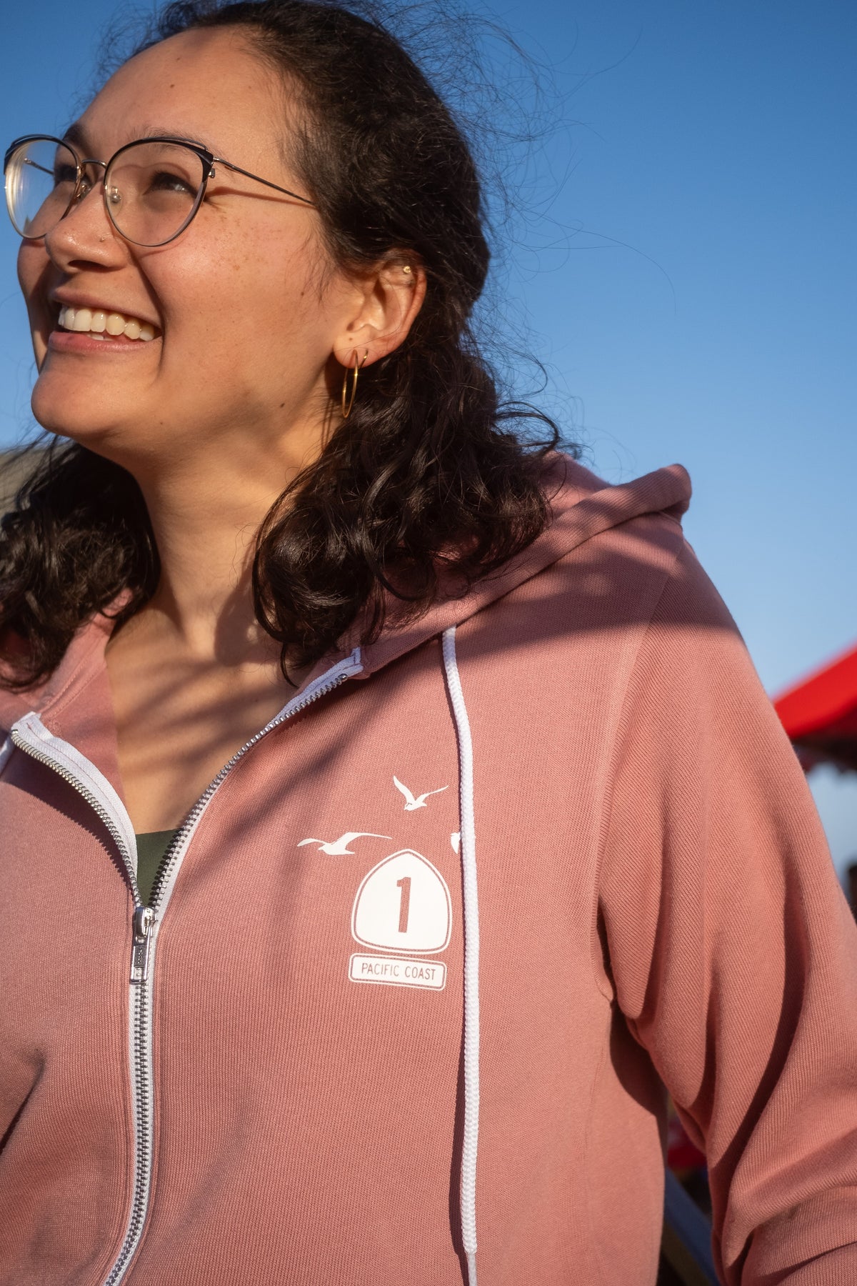 A woman wearing round glasses and a dusty rose zip-up hoodie with white drawstrings and a small white graphic on the left chest, photographed outdoors against a clear blue sky.