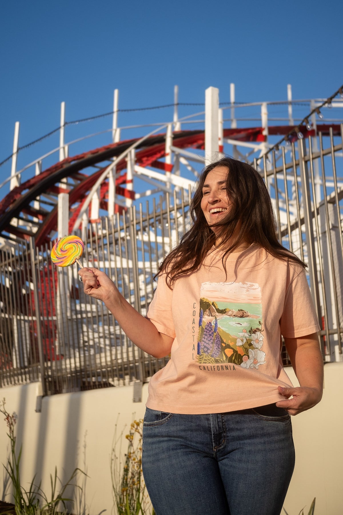 A woman wearing a peach graphic t-shirt and blue jeans stands outdoors in front of a roller coaster, holding a yellow and pink swirl lollipop.