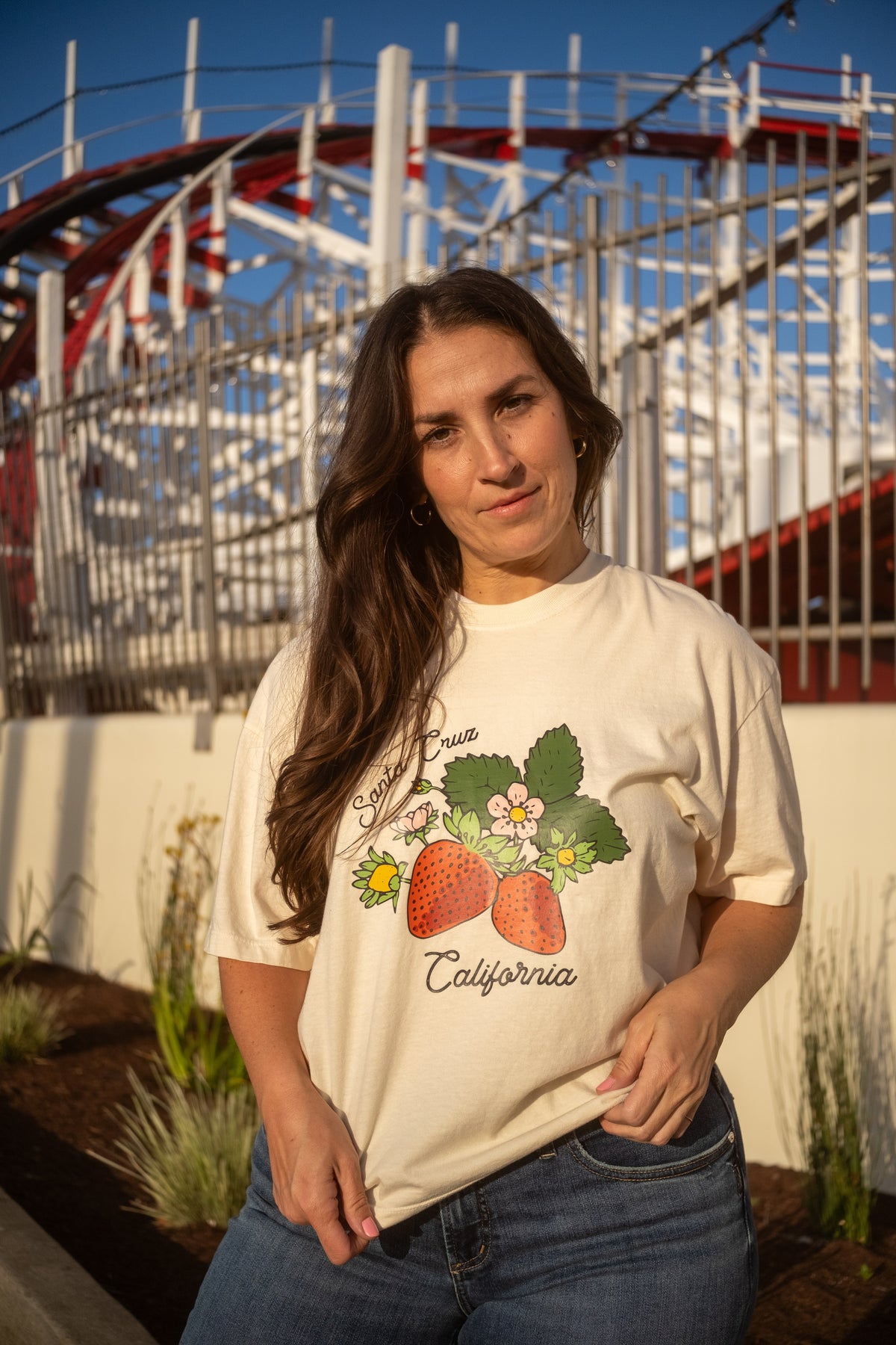 A woman wearing a cream-colored graphic t-shirt with a strawberry and 'California' print, standing outdoors in front of a white fence and red roller coaster.