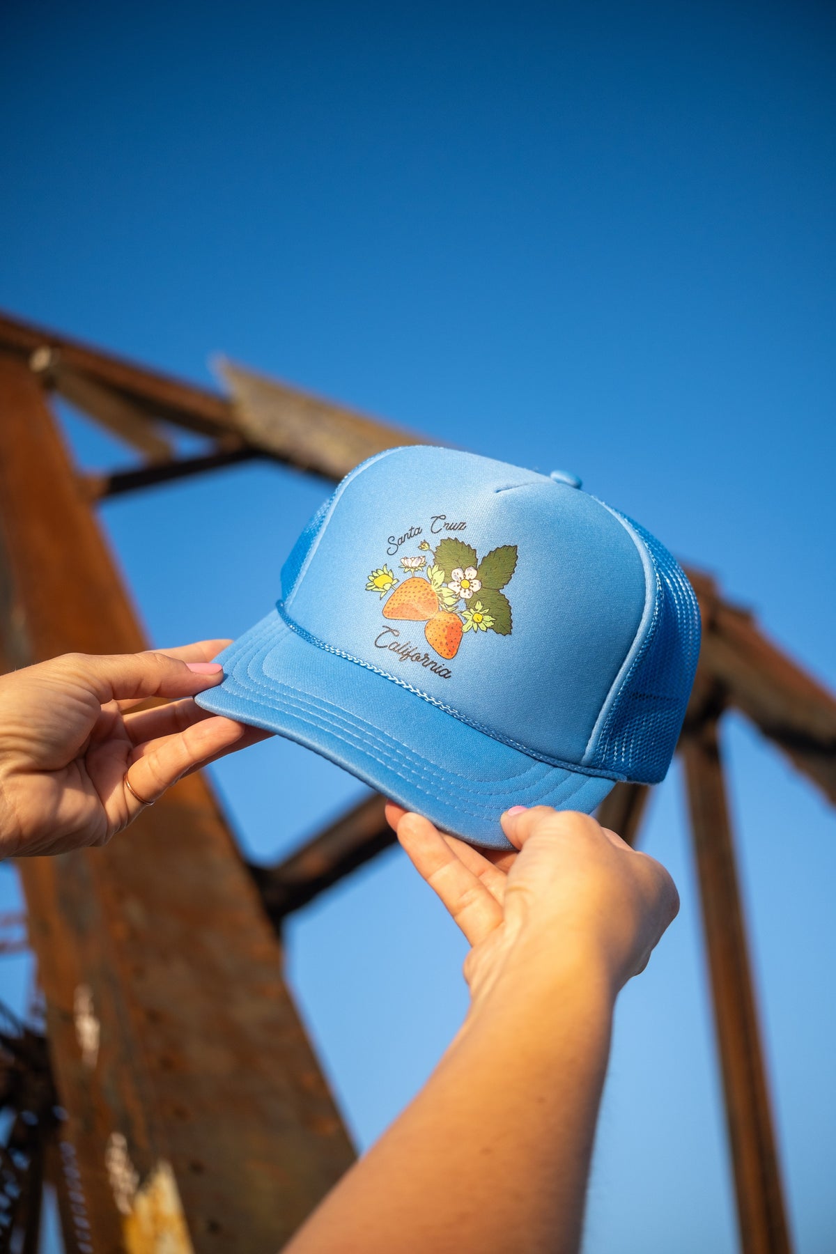 A light blue trucker hat with a 'Santa Cruz California' graphic, held by two hands against a bright blue sky and rusty metal structure.