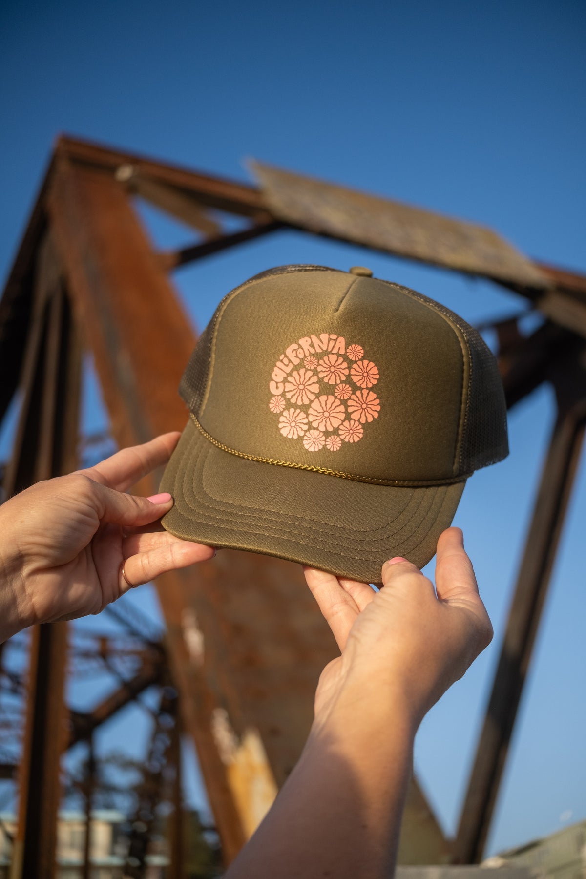 An olive green trucker hat with a pink floral pattern on the front, held by two hands against a blurred rusty metal structure and blue sky background.