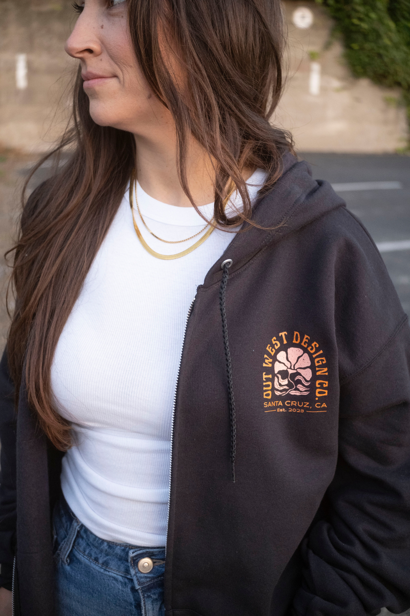 Woman wearing a black hoodie with a logo and text, standing outdoors.