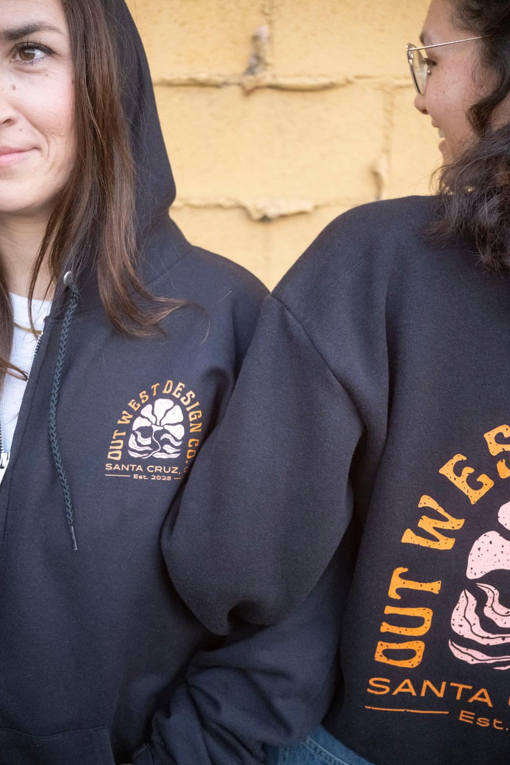 Two people wearing black hoodies with visible branding against a textured wall.