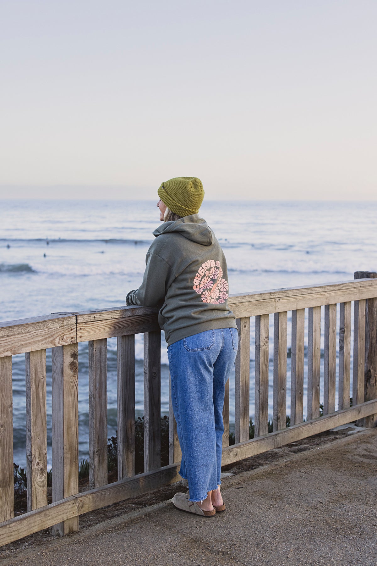 Person wearing a green beanie and hoodie with a floral design, standing on a wooden deck overlooking the ocean.