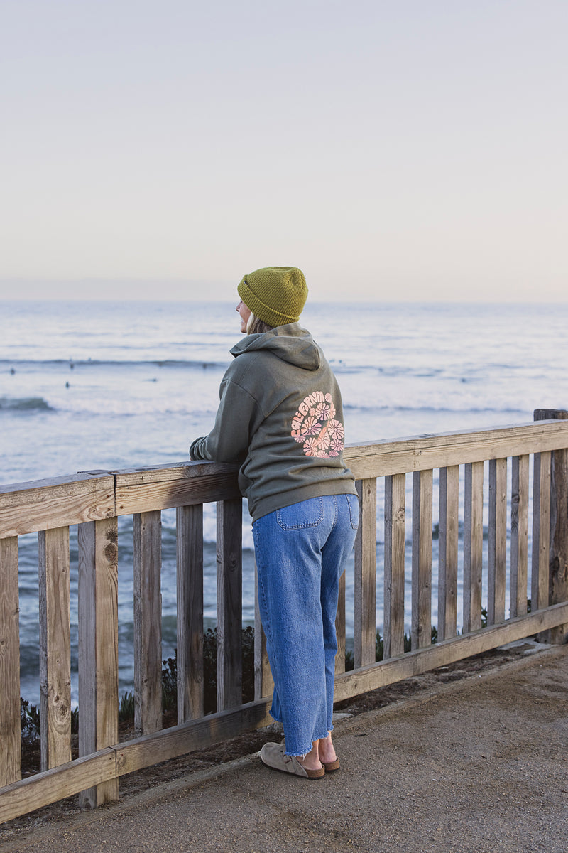 Person wearing a green beanie and hoodie with a floral design, standing on a wooden deck overlooking the ocean.