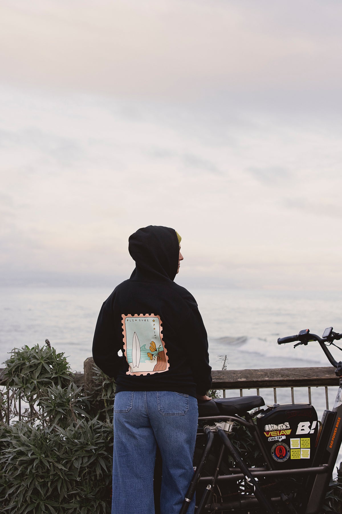 A woman in a black hoodie standing next to a bike and looking out over the ocean. 