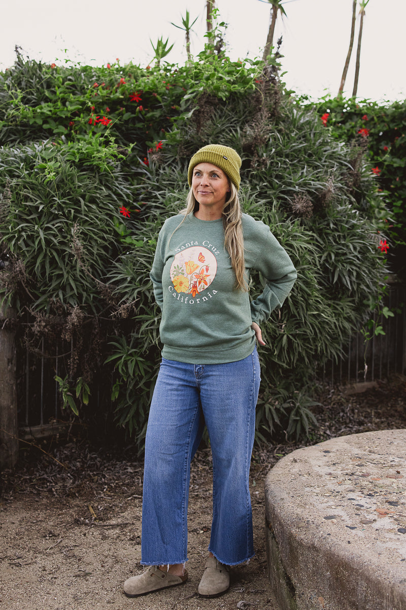 Woman wearing a green sweatshirt with a graphic design and blue jeans standing in front of a hedge.