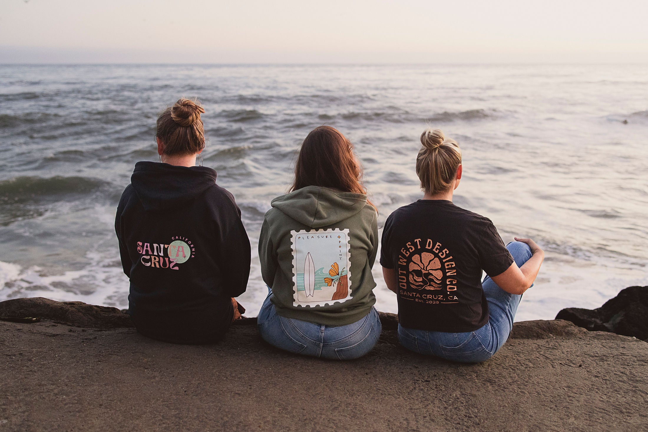 Three people sitting on a beach wearing hoodies with visible text and graphics.