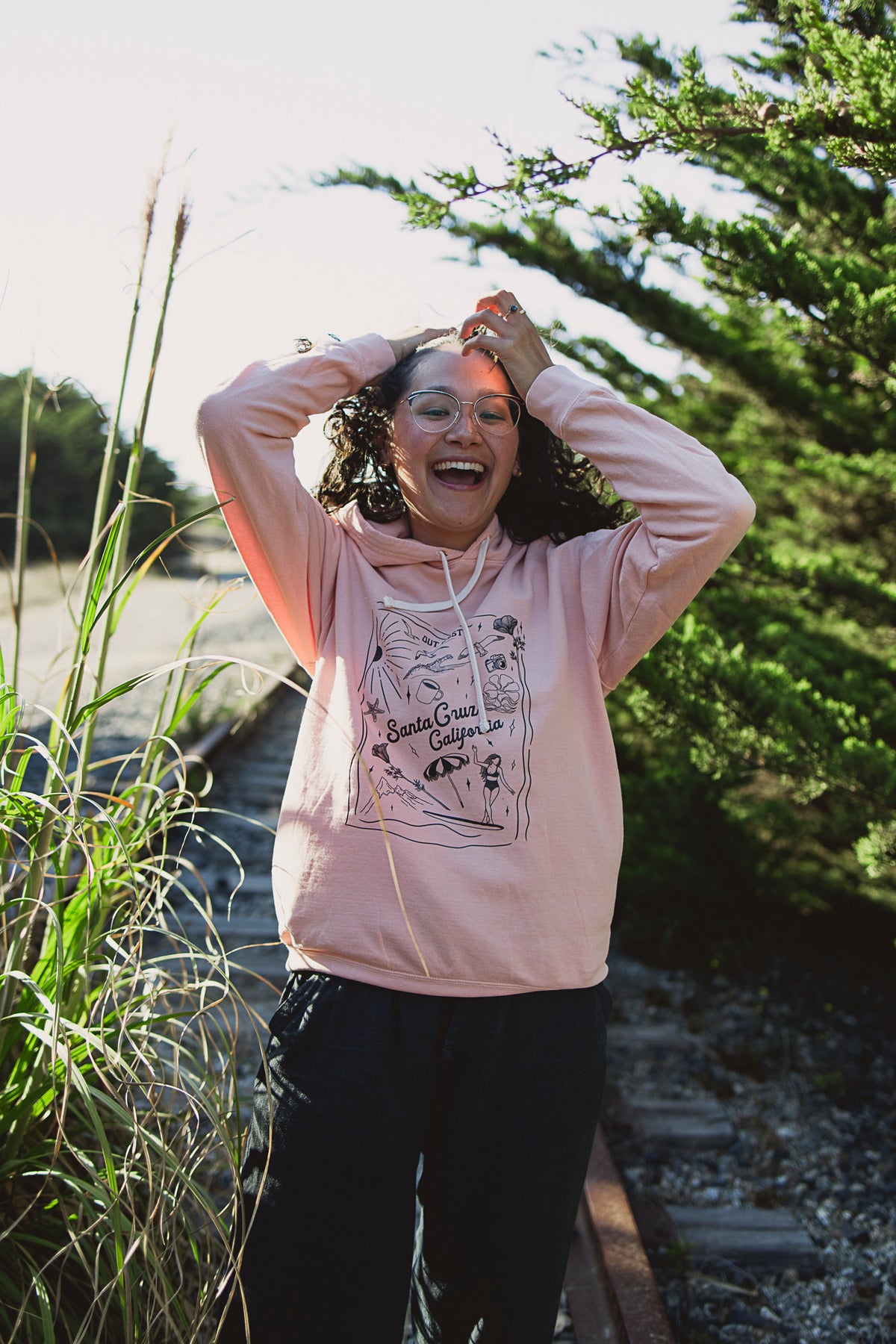 Person wearing a pink hoodie with a graphic design, standing outdoors near tall grass and trees.