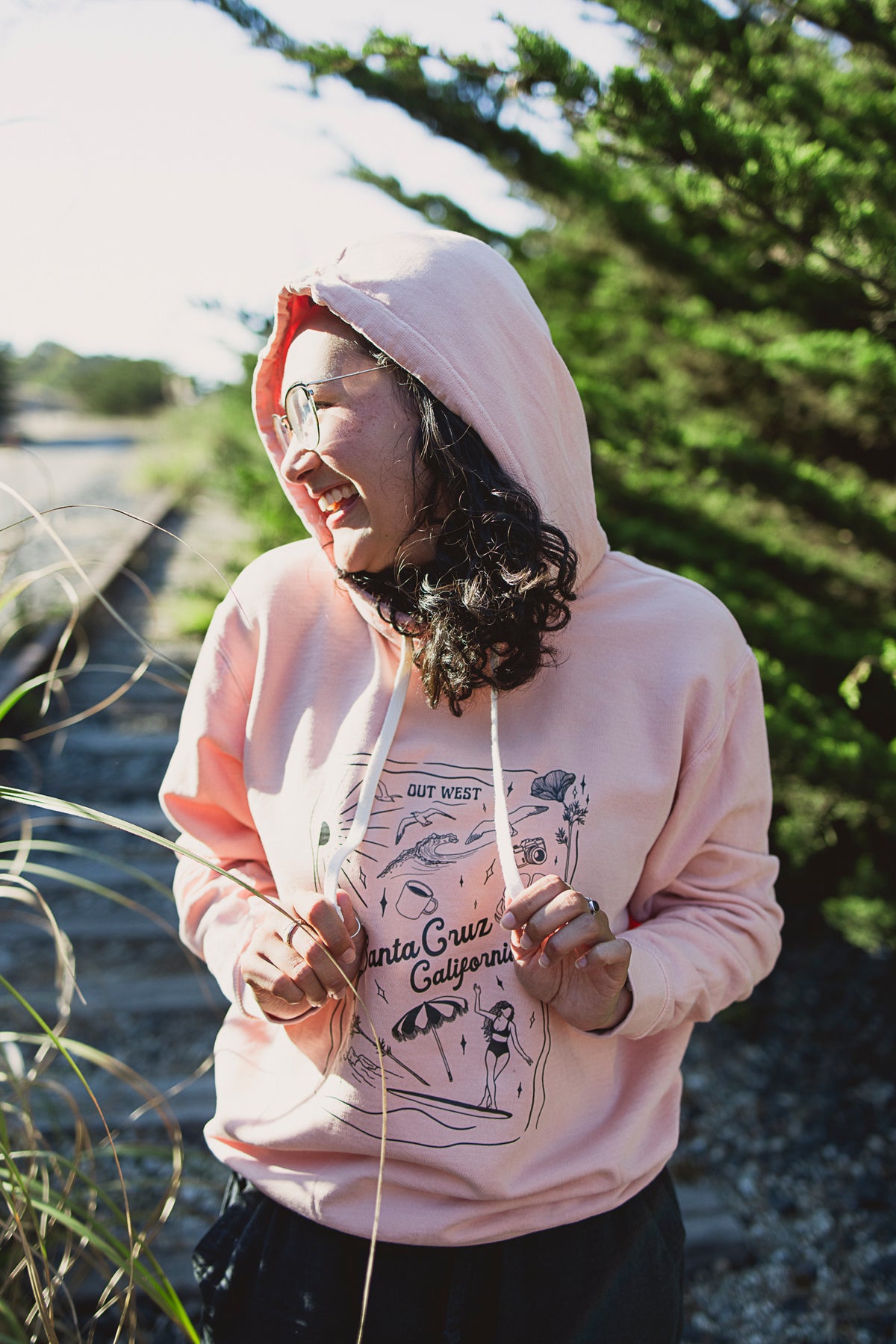 Person wearing a pink hoodie with a graphic design, standing outdoors near greenery.